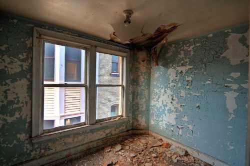 An empty, dilapidated room with peeling blue paint, cracked walls, and a damaged ceiling, featuring two windows.