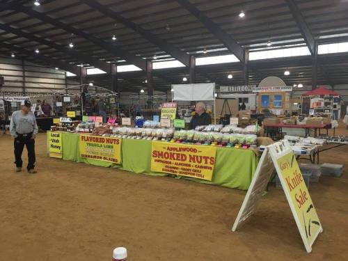 A vendor booth at a market featuring flavored nuts, smoked nuts, and a sign for a knife sale in a spacious indoor setting.