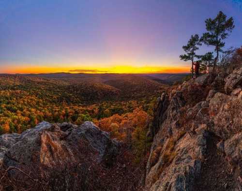 A panoramic view of a colorful sunset over a valley, framed by rocky cliffs and trees.