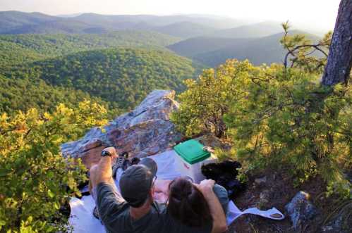 A couple sits on a rocky ledge, enjoying a scenic view of rolling green hills at sunset.