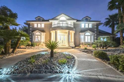 A grand, illuminated mansion with a circular driveway, surrounded by palm trees and landscaped rocks at dusk.