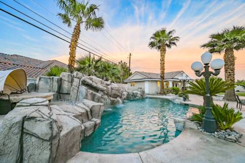 A serene pool area with rock formations, palm trees, and a sunset sky in the background.