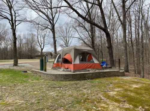 A tent set up on a stone platform in a wooded area, surrounded by bare trees and a grassy field.