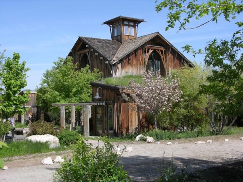 A rustic wooden building surrounded by greenery and blooming trees under a clear blue sky.