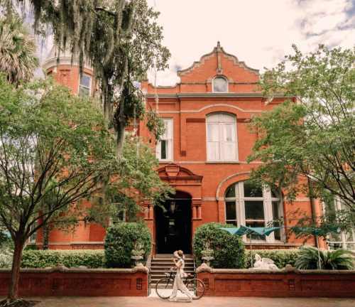 A woman walks with a bicycle in front of a historic red brick building surrounded by greenery.