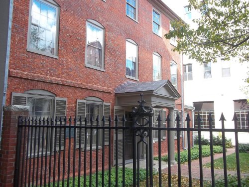 A brick building with large windows, a decorative entrance, and a wrought iron fence, surrounded by greenery.