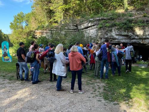 A group of people gathered outside a cave entrance, surrounded by greenery and a clear blue sky.
