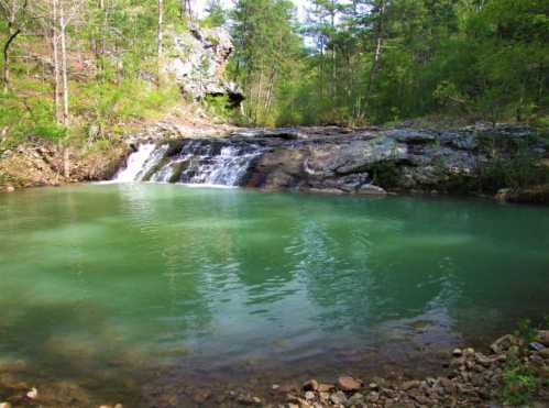 A serene turquoise pool with a small waterfall, surrounded by lush green trees and rocky terrain.