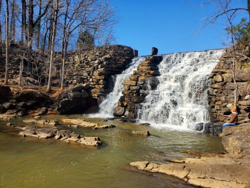 A serene waterfall cascades over stone steps into a calm river, surrounded by trees under a clear blue sky.