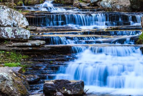 A serene waterfall cascades over rocky steps, surrounded by lush greenery and smooth stones.