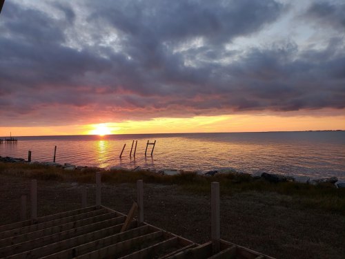 A serene sunset over calm waters, with clouds reflecting warm colors and silhouettes of old posts along the shore.