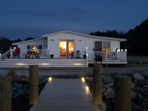 A cozy white house at dusk, with a deck, lights, and a wooden dock leading to rocky shore.