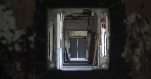 A view through a square opening, revealing a long, dimly lit corridor with doors at the end.