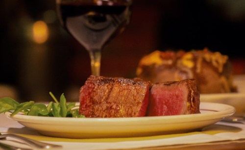 A plate with two pieces of cooked steak, garnished with greens, and a glass of red wine in the background.