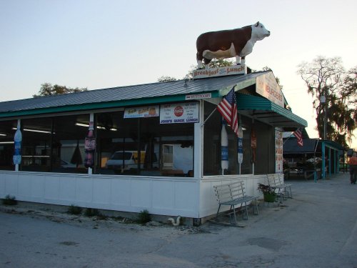 A rustic diner with a cow statue on the roof, featuring outdoor seating and American flags.
