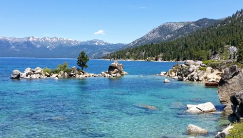 A serene lake scene with clear blue water, rocky shores, and mountains in the background under a bright blue sky.