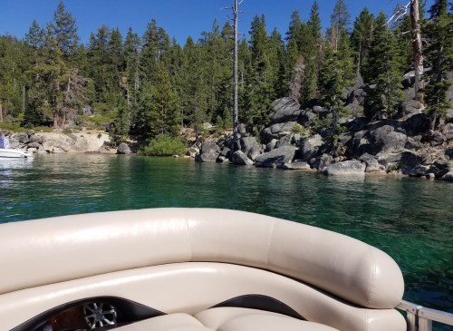 A view from a boat showing clear green water, rocky shoreline, and lush trees under a blue sky.