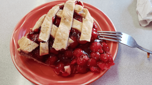 A slice of cherry pie on a red plate, topped with a lattice crust and surrounded by glossy cherries. A fork rests beside it.
