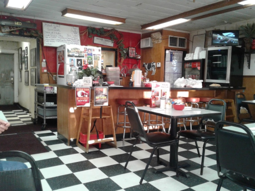A cozy diner interior with checkered floors, a counter, and tables, featuring a menu board and food displays.