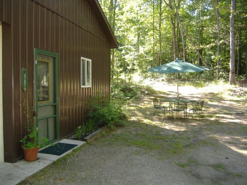 A rustic brown building with a green door, surrounded by trees, and a patio set with an umbrella nearby.
