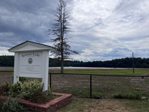 Sign for Kenoza Lake with a view of the lake and cloudy sky in the background, surrounded by a fence and greenery.