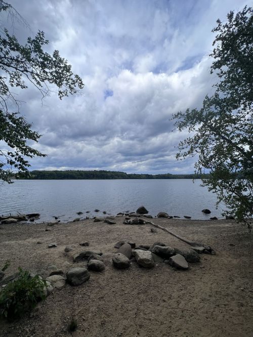 A serene lakeside view with rocky shorelines, surrounded by trees and a cloudy sky.
