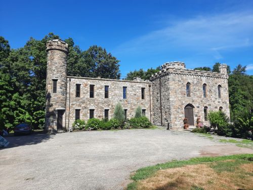 A stone castle with towers, surrounded by greenery and a gravel driveway under a clear blue sky.