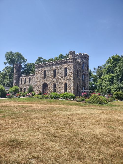 A stone castle with a tower, surrounded by greenery and colorful flowers, under a clear blue sky.