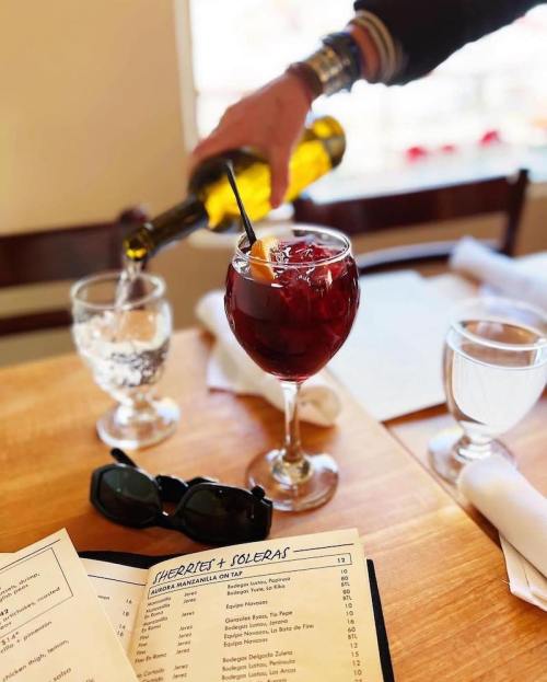 A person pours a drink into a glass of red liquid, with a menu and sunglasses on a wooden table.