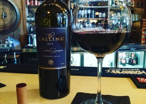 A bottle of 2013 The Calling Cabernet Sauvignon next to a glass of red wine on a bar counter.