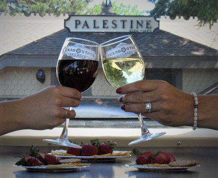 Two hands clink wine glasses filled with red and white wine, surrounded by plates of strawberries and crackers.