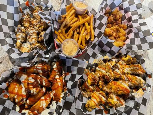 A spread of various chicken wings, sweet potato fries, and fried bites in checkered paper baskets with dipping sauces.