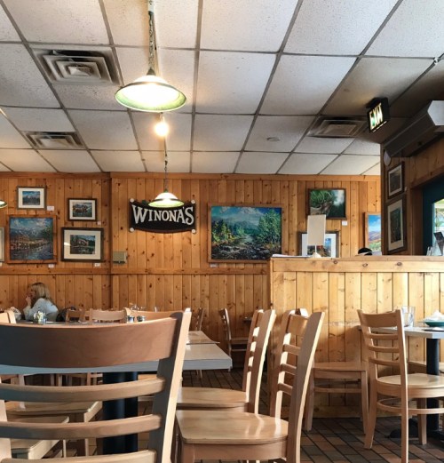 Cozy restaurant interior with wooden walls, tables, and framed landscape paintings. A sign reads "Winona's."
