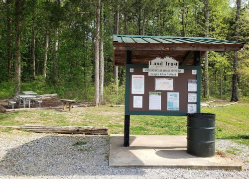 A wooden kiosk with a green roof displaying information about the Land Trust, surrounded by trees and a picnic area.