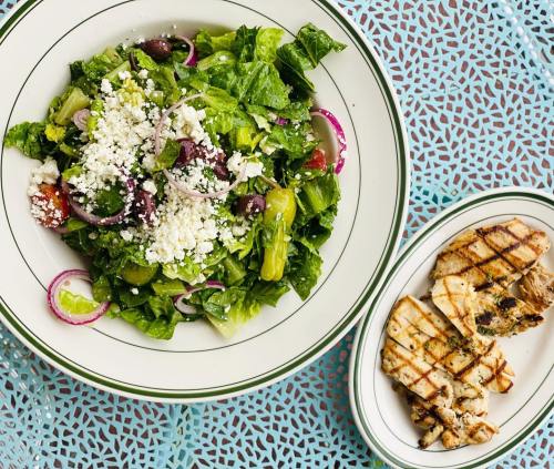A fresh salad with greens, olives, and feta cheese, served alongside grilled chicken on a decorative table.