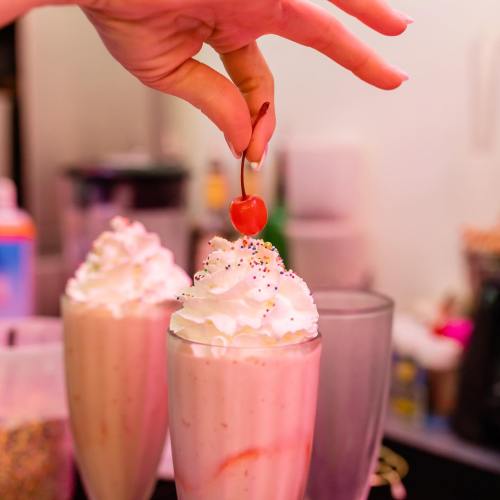 A hand places a cherry on top of a milkshake with whipped cream and colorful sprinkles.