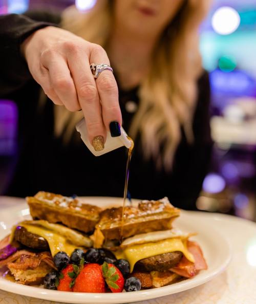 A hand pours syrup over a plate of waffles, bacon, and fresh berries.