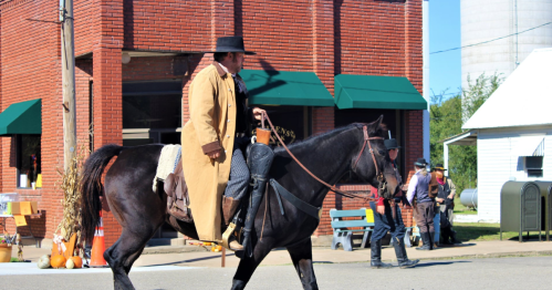 a man in a long coat and a cowboy hat sits on a black horse and rides down the road during hte woodibine kansas fall festival on a sunny day