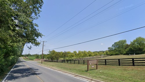 A rural road lined with trees and a wooden fence, leading to a grassy field under a clear blue sky.