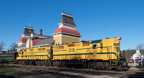 Two yellow and green locomotives parked beside a historic train station under a clear blue sky.