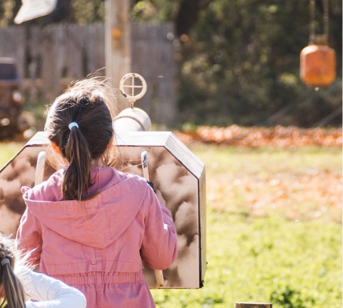 A child in a pink jacket looks through a telescope in a sunny outdoor setting with trees and leaves in the background.