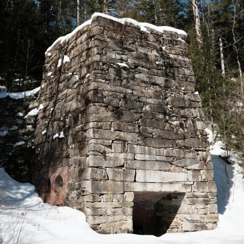 A tall, weathered stone structure partially covered in snow, surrounded by trees in a winter landscape.