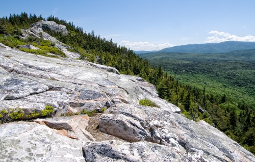 A rocky outcrop overlooks a lush green valley and distant mountains under a clear blue sky.