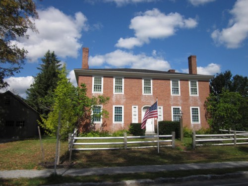 A brick house with white trim and an American flag, surrounded by trees and a wooden fence under a blue sky.