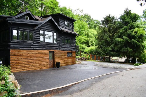 A black log cabin with wooden accents, surrounded by lush greenery and a paved driveway.
