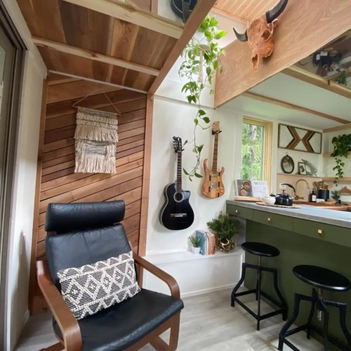 Cozy interior with a black chair, wooden accents, plants, and a kitchen area featuring green cabinets and decor.