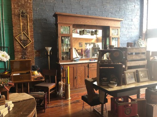 A vintage shop interior featuring an antique cabinet, wooden furniture, and various collectibles against a brick wall.