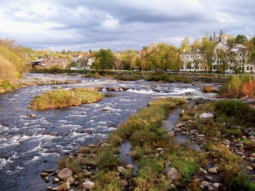 A scenic river flows through a landscape with greenery and buildings under a cloudy sky.