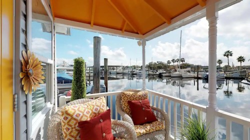 Cozy porch with wicker chairs overlooking a marina filled with boats and palm trees under a bright blue sky.