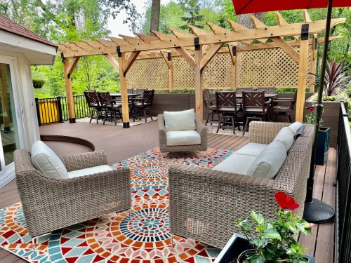 A cozy outdoor seating area with wicker furniture, a colorful rug, and a wooden pergola surrounded by greenery.